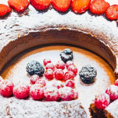 A front view of a Gluten Free Marble Cake, topped with icing sugar, sliced strawberries and other types of fruits
